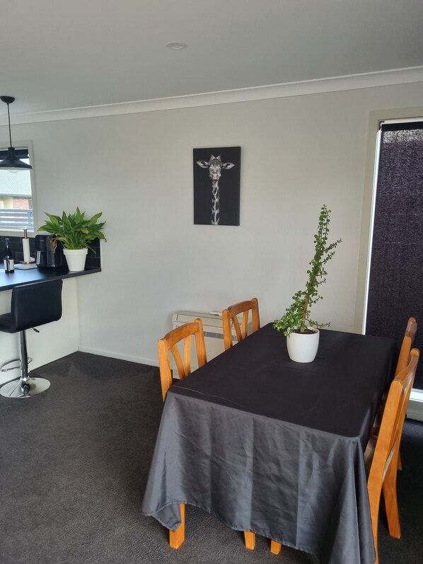 A dining area with a table covered by a black tablecloth, surrounded by wooden chairs. There is a potted plant on the table and another on the wall. A black artwork of a giraffe is hanging on the wall.