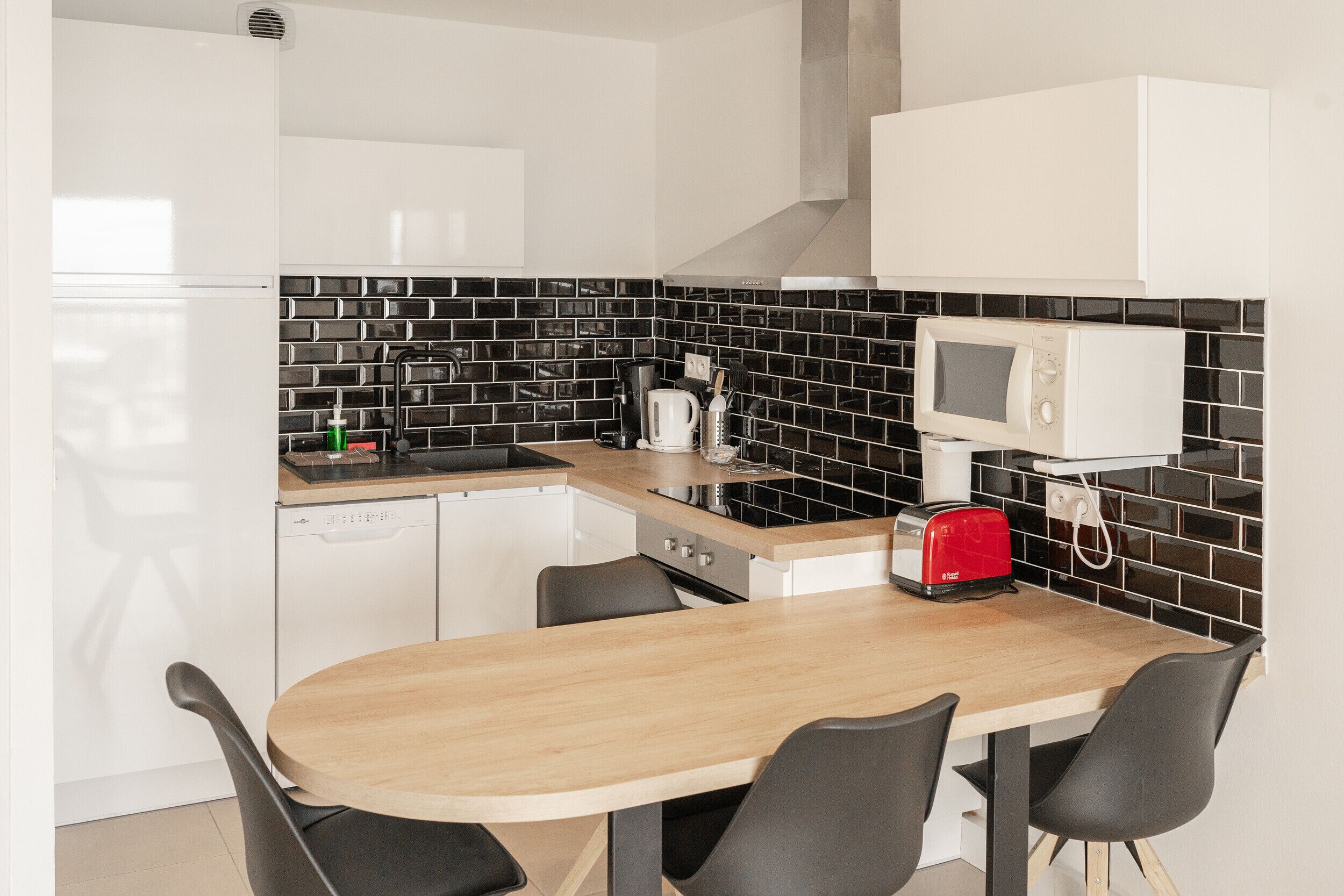 Una cocina moderna con una mesa de madera y sillas negras, un refrigerador blanco, un microondas y una cafetera.