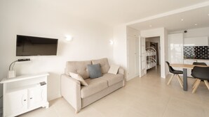 Una sala de estar moderna con un sofá, un televisor montado en la pared, una zona de comedor con mesa y sillas, y una cocina visible a través de un vano abierto.
