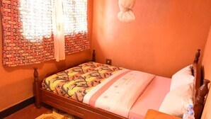 A single bed with a patterned bedspread, a bedside table with a water bottle, and a window with patterned curtains.