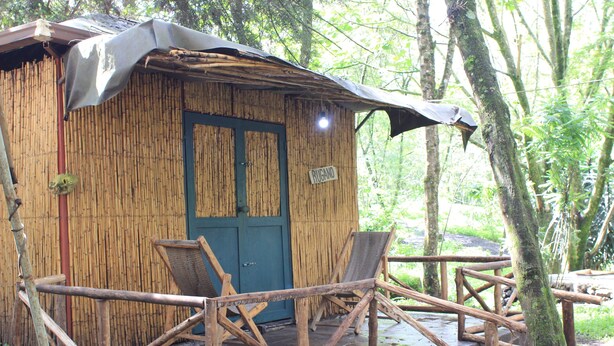 Une hutte rustique en bambou avec une porte bleue, un petit porche doté de balustrades en bois et une enseigne sur laquelle on peut lire « HUGO ».