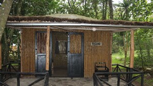 Une cabane rustique en bois, avec une façade en bambou, une enseigne indiquant « RUGANO » et une zone d’entrée pavée de pierres, agrémentée d’un banc.