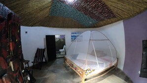 A room with a bed under a mosquito net, a chair, a small table, and a door leading to another room. - Red Rocks Rwanda - Campsite & Guesthouse (Nyakinama)