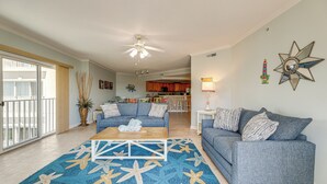 Ein geräumiges Wohnzimmer mit einem blauen Teppich mit Sternfischmuster, einem Holztisch und einem grauen Sofa. An der Wand befinden sich ein Deckenventilator und ein sternförmiger Spiegel.