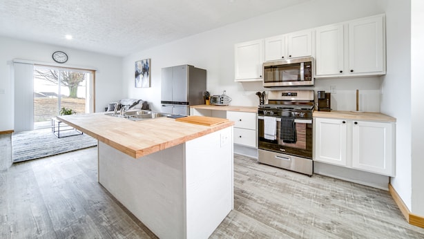 Une cuisine moderne avec des meubles blancs, un îlot central en bois, des appareils électroménagers en acier inoxydable et une grande fenêtre.