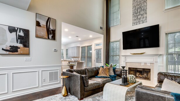 A spacious living room with a fireplace, a large flat-screen TV mounted above it, and a modern kitchen in the background. - Modern Stone Mountain Home w/ Spacious Deck, Grill (Stone Mountain)