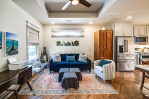 A modern living room with a dark blue sofa, a white armchair, and a coffee table. There is a kitchen area with stainless steel appliances and wooden cabinets. - Walkable Dallas Bungalow: 4 Mi to Downtown (Dallas)