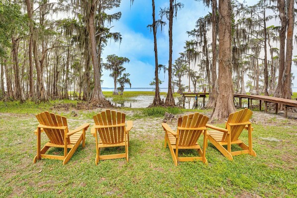 Um cenário de praia com quatro cadeiras de madeira Adirondack dispostas em semicírculo, voltadas para um corpo d'água, rodeadas por árvores altas com barba-de-mosmo.