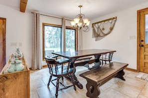 Uma sala de jantar com mesa de madeira, cadeiras, um banco, um candelabro e uma janela com cortinas.