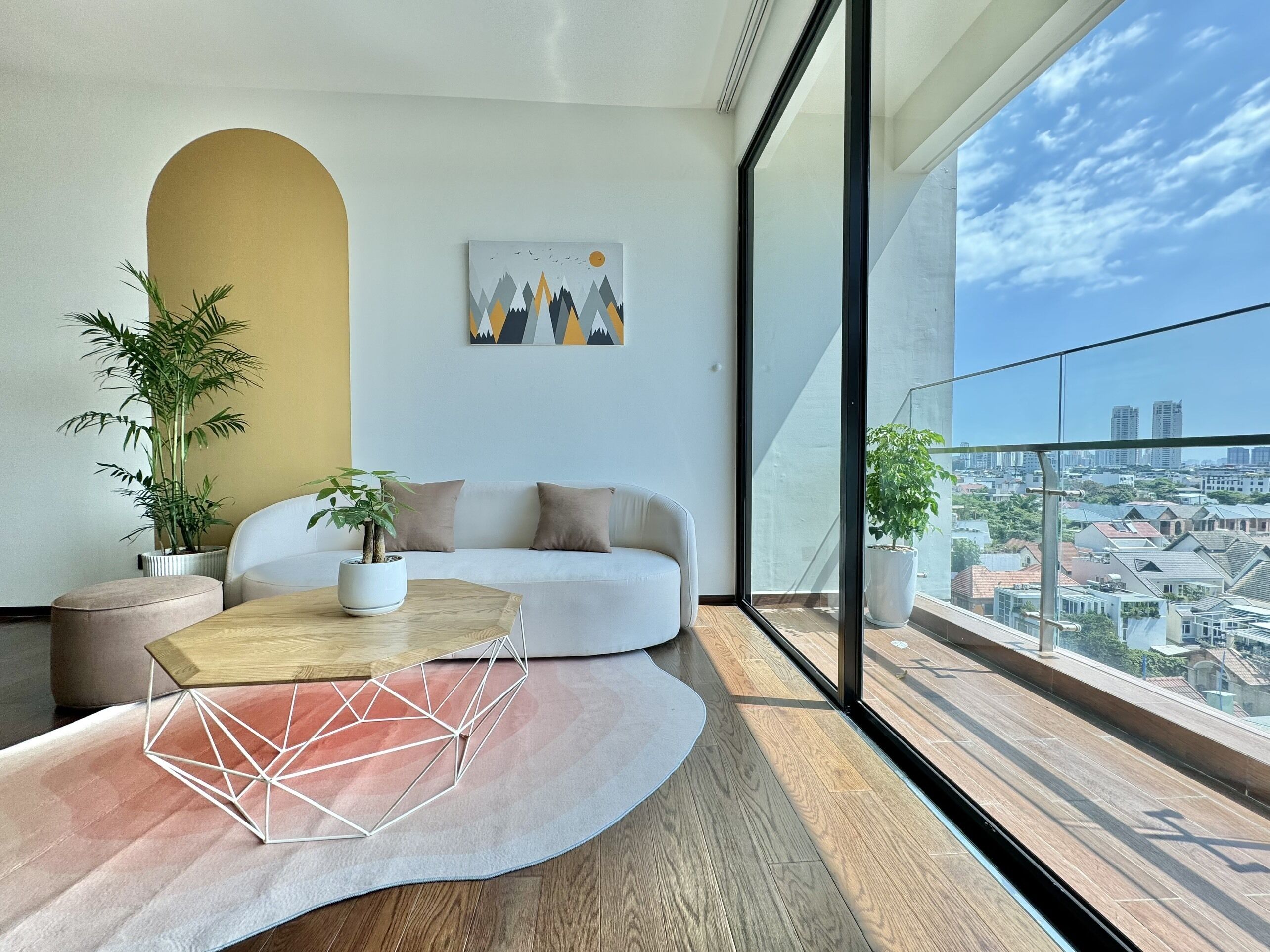 Un salon moderne avec un canapé blanc, une table basse aux motifs géométriques et deux plantes en pot. Un tableau représentant des montagnes et des oiseaux est accroché au mur.
