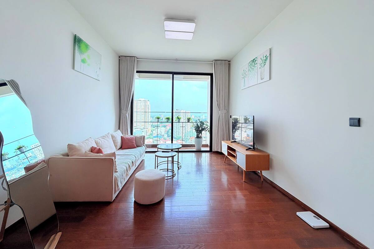 A modern living room with a sofa, ottoman, TV stand, and a large window with a city view.