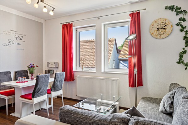 A modern living room with a dining area, large windows with red curtains, a clock on the wall, and a radiator.