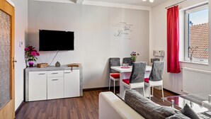 Un salon moderne doté d’un téléviseur à écran plat, d’un coin repas avec des chaises rouges, d’un canapé et d’une fenêtre ornée de rideaux rouges.