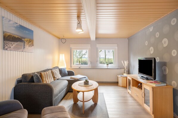 A modern living room with a sofa, a round coffee table, a TV on a wooden stand, and a window with a view of trees.
