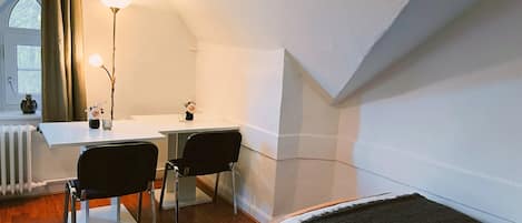 A room with a desk, two chairs, a bed, a radiator, and a window with curtains.