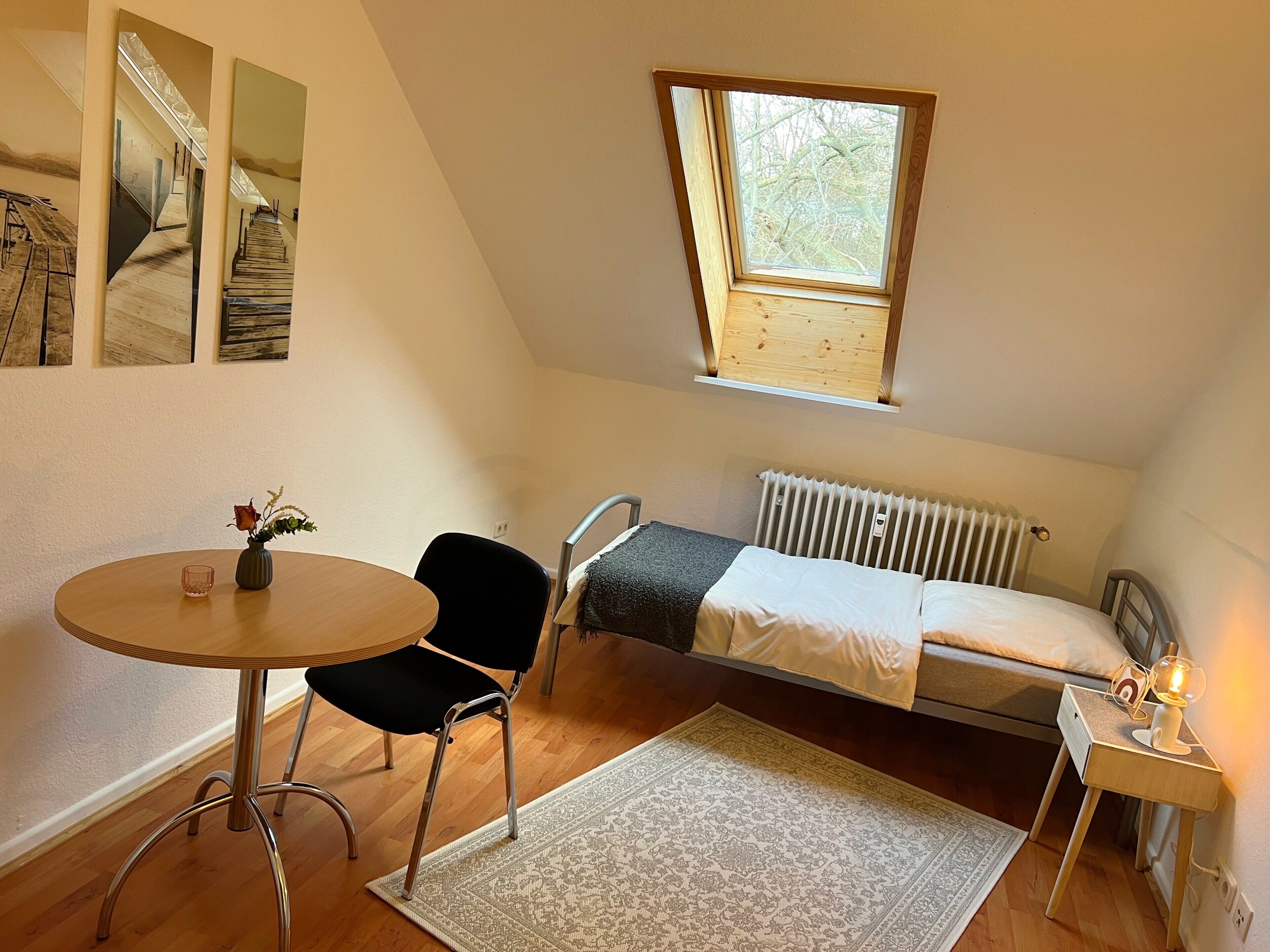 A small, well-lit room with a single bed, a table with a chair, a radiator, and a window.