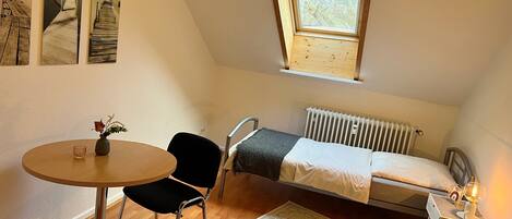 A small, well-lit room with a single bed, a table with a chair, a radiator, and a window.