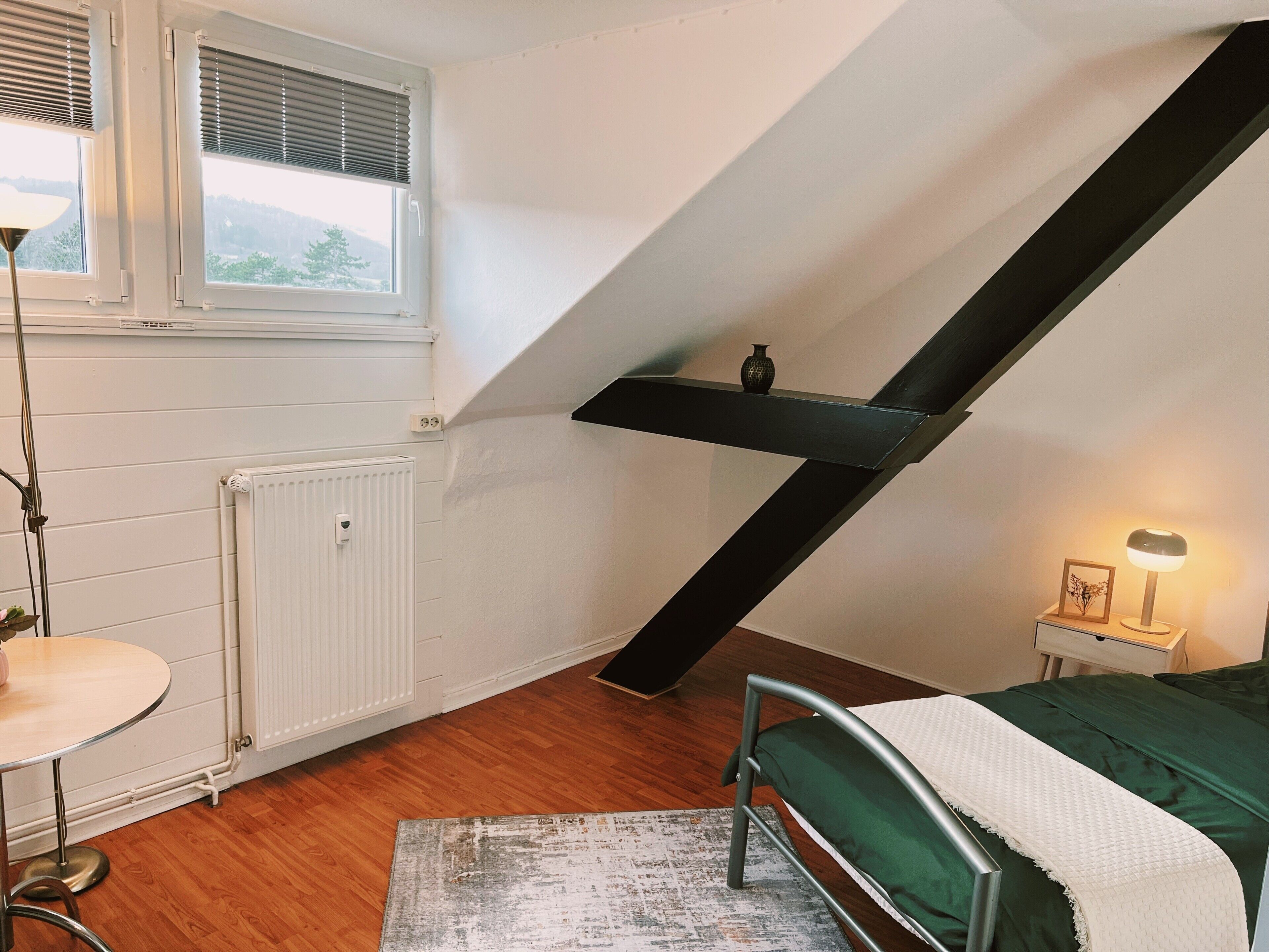 Una camera da letto con soffitto spiovente, un letto, un comodino, una lampada e un termosifone.