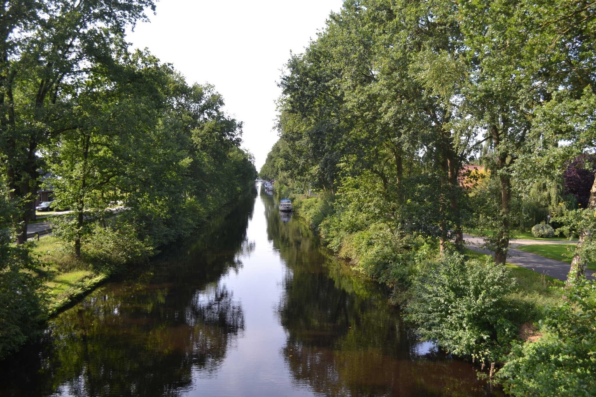En smal kanal omgivet af tæt bevoksning, med en vej på den ene side og en grøn græsplæne på den anden.