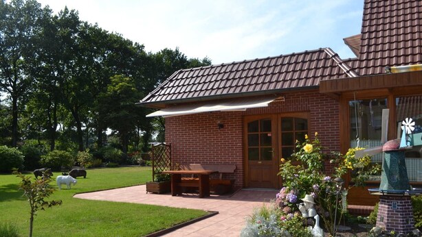 A brick house with a tiled roof, a patio with a table and chairs, and a garden with various plants and a windmill.