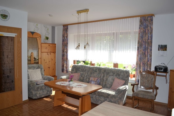 Ein Wohnzimmer mit einer Couch, Sesseln, einem Holztisch, einem Esstisch und einem Fenster mit Vorhängen.