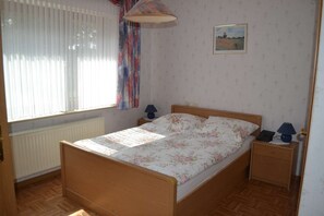 Un dormitorio con cama de madera, una mesita de noche con lámpara, una ventana con cortinas y un cuadro enmarcado en la pared.