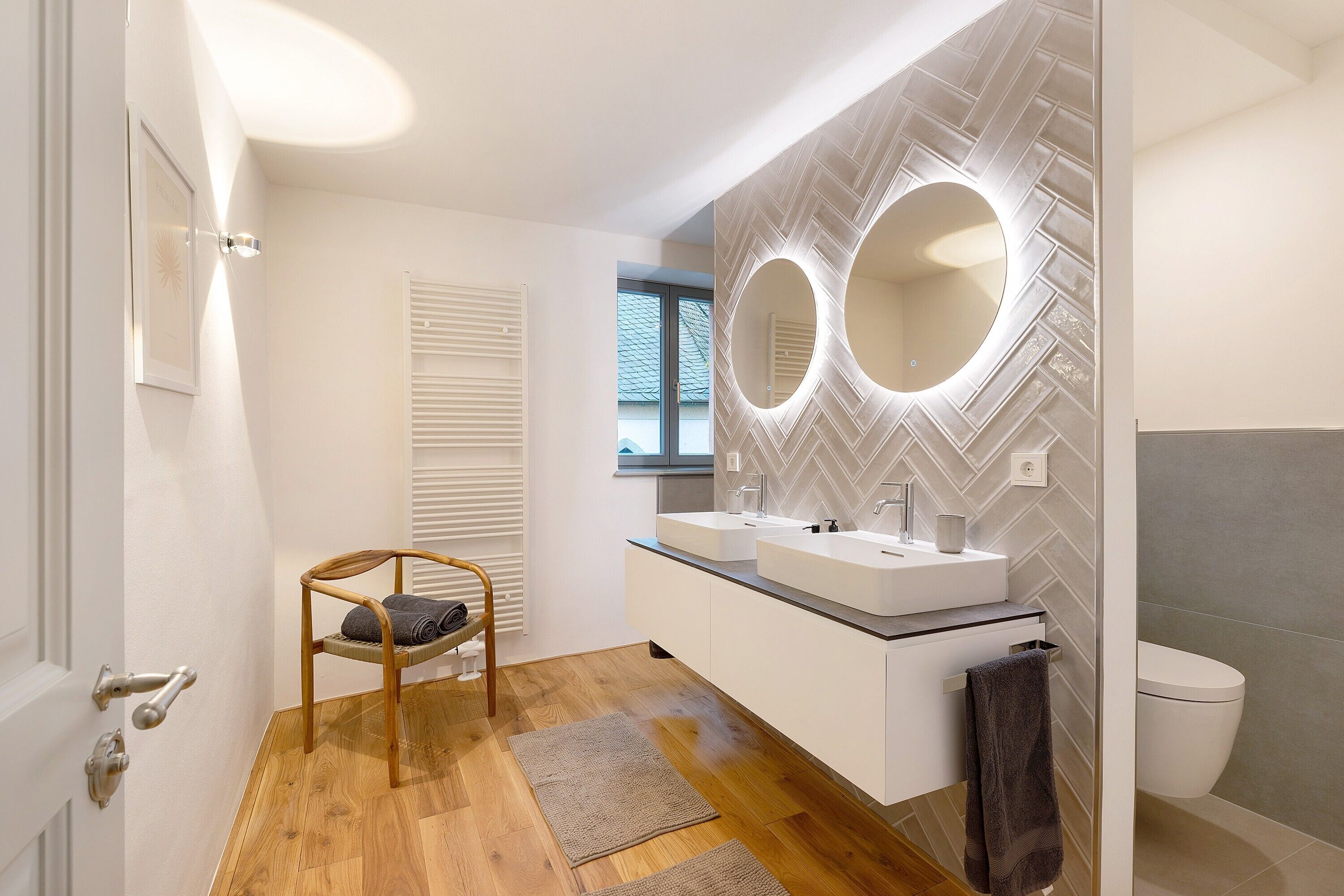 Une salle de bain moderne avec un plancher en bois, un meuble-lavabo blanc doté de deux vasques, un grand miroir et des toilettes.