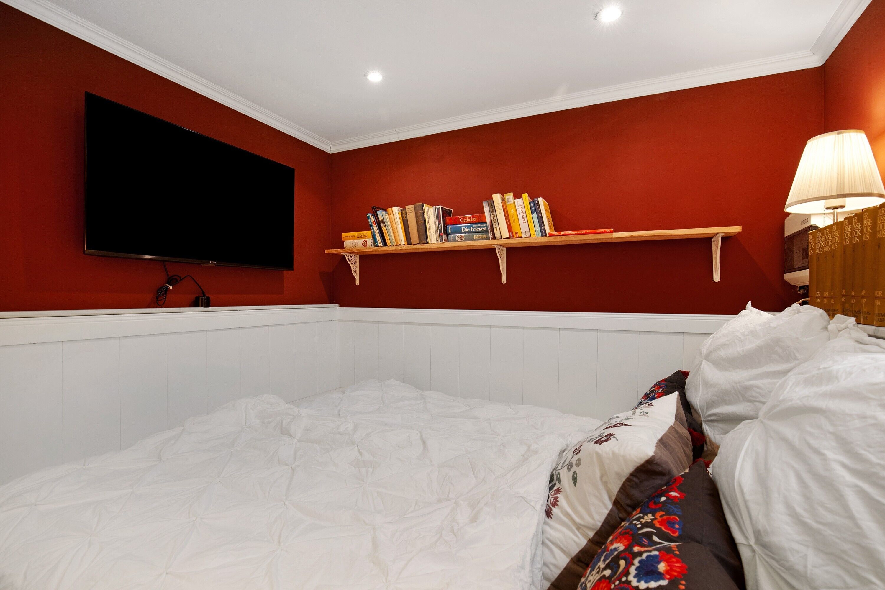 A bedroom with a bed, a mounted TV, a shelf with books, and a bedside lamp.