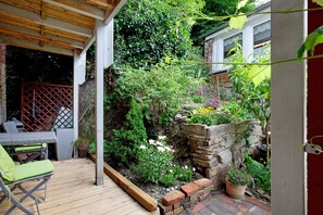 Um deck de madeira com mesa e cadeiras, cercado por um jardim com diversas plantas e um muro de pedra.