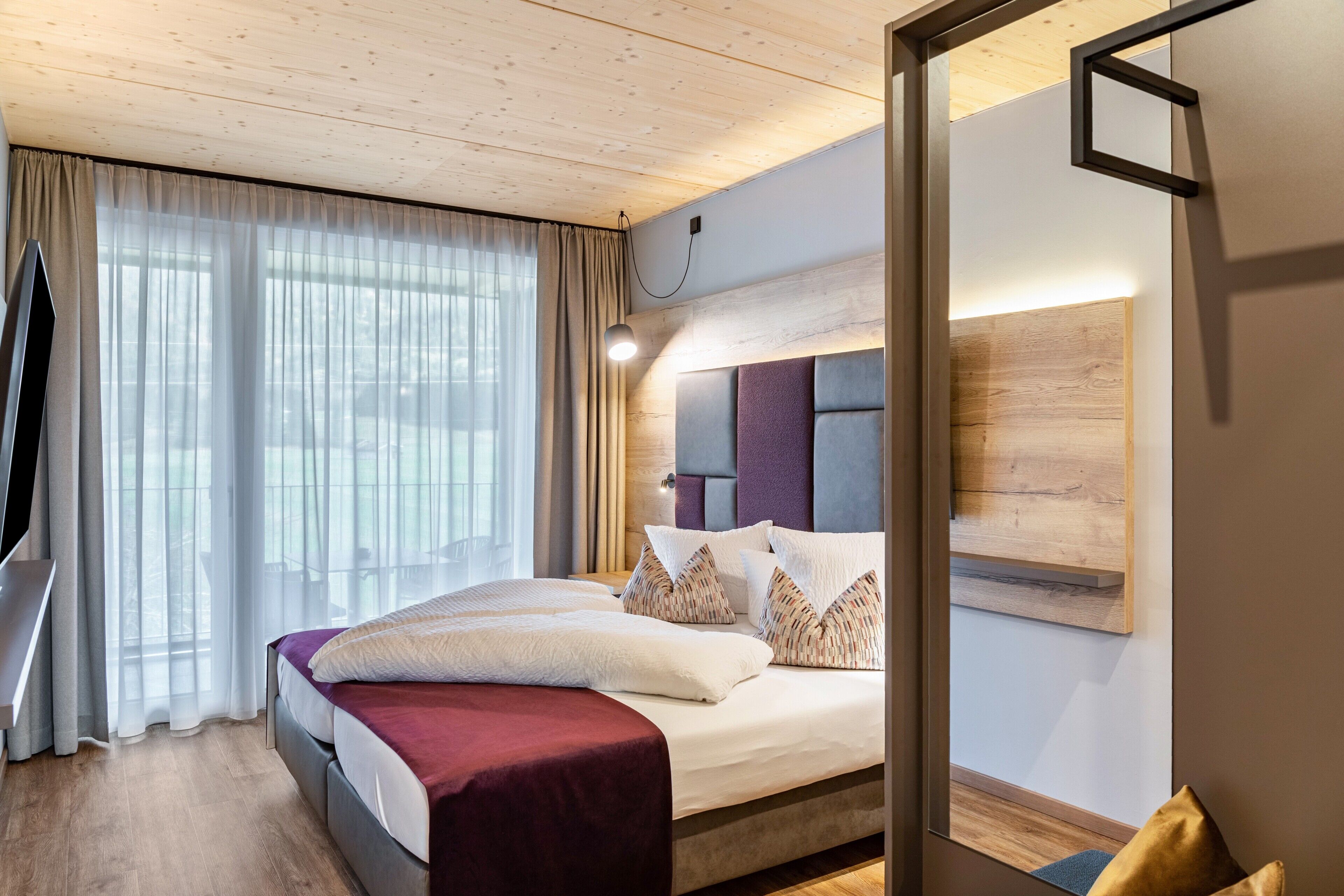 Une chambre d’hôtel moderne avec un grand lit, une télévision, un plafond en bois et une grande fenêtre avec des rideaux.