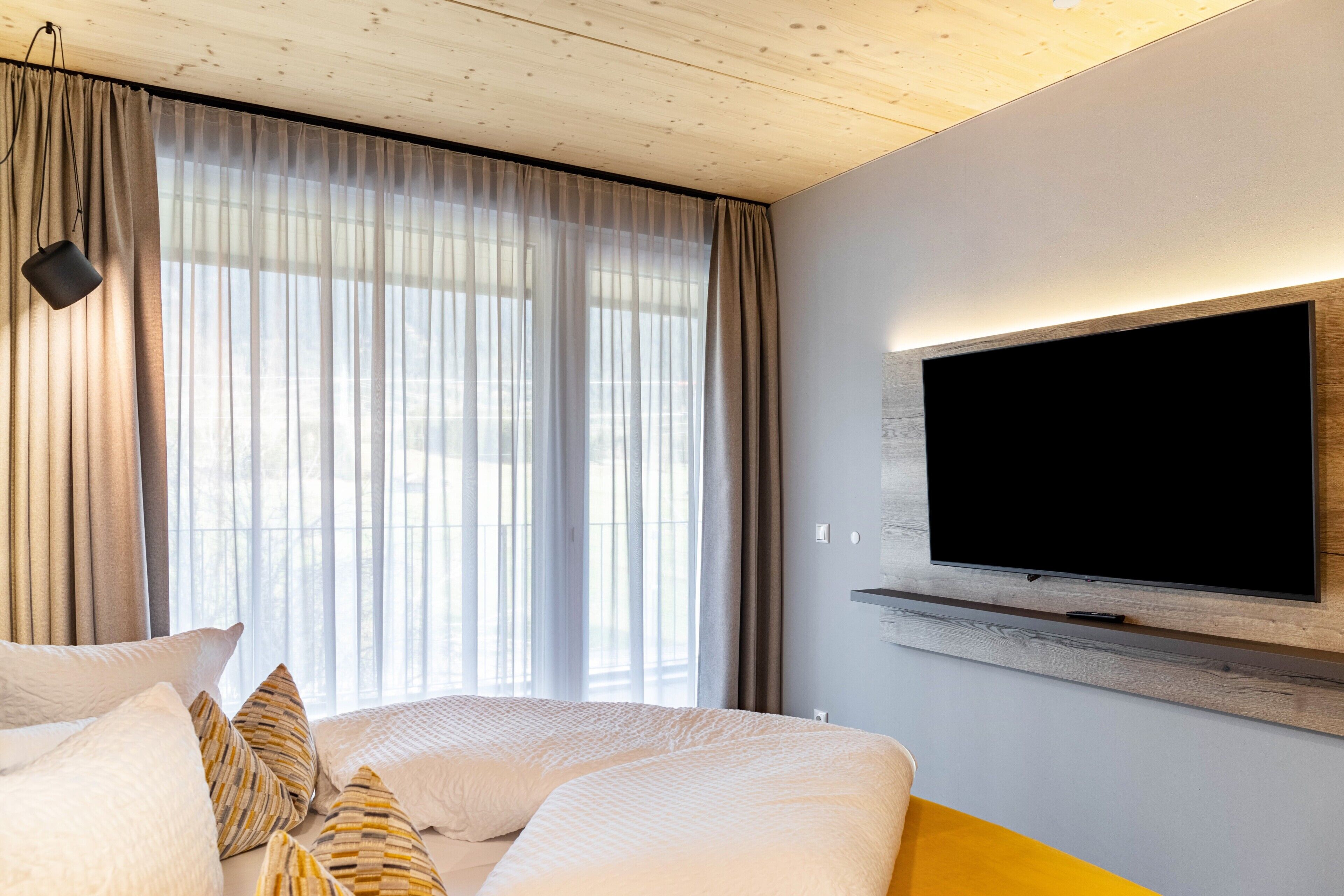 A modern hotel room with a large bed, a television mounted on the wall, and a window with sheer curtains.