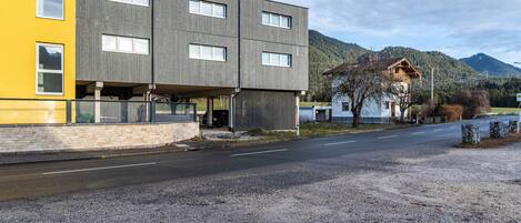 A modern building with a yellow facade and a grey upper section, a gravel driveway, and a mountain in the background.