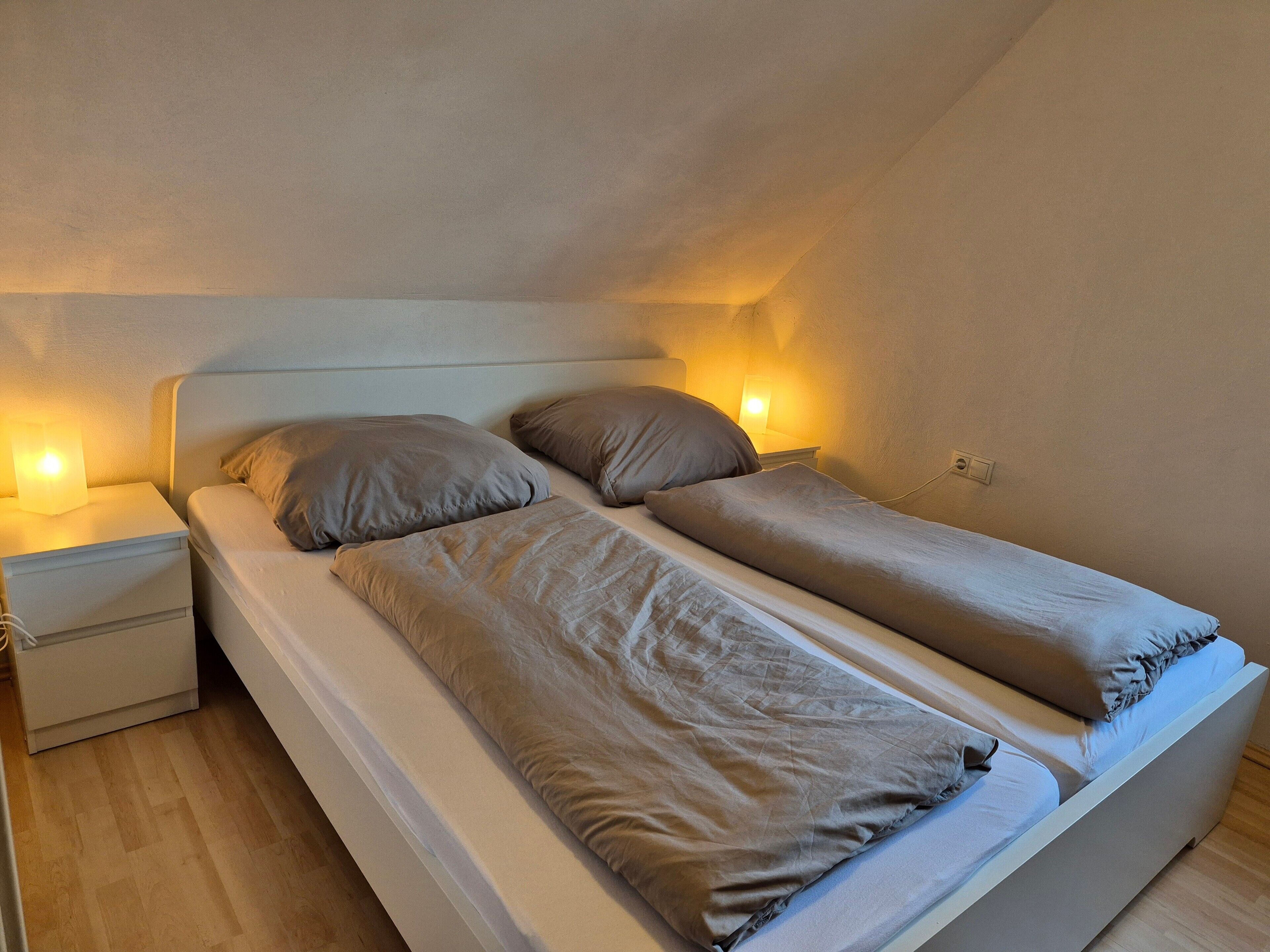 A twin bed with two pillows, a bedside table with a lamp, and a power outlet on the wall.