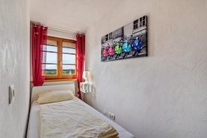 Une chambre à coucher avec un lit, une fenêtre ornée de rideaux rouges, une lampe fixée au mur et un tableau représentant des scooters.