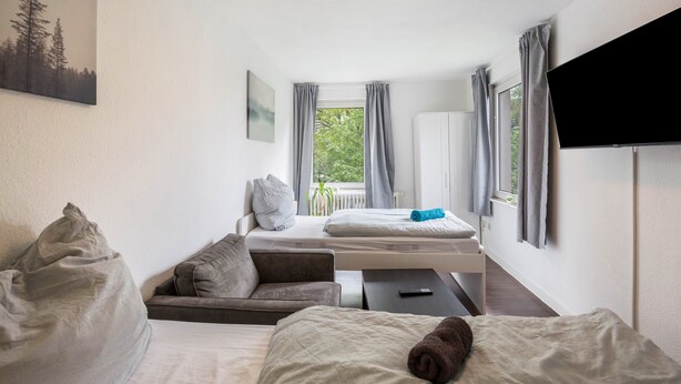 Quarto de hotel com cama, sofá, mesinha, televisão e janela com cortinas.