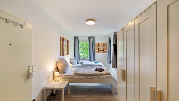 Ein Schlafzimmer mit zwei Betten, einem Nachttisch, einer Lampe, einem Fenster mit Vorhängen und einem an der Wand montierten Fernseher.