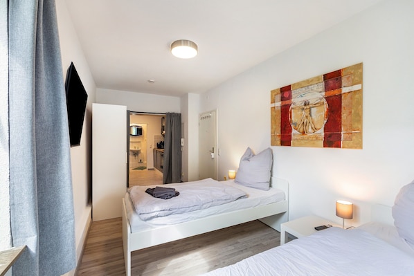 A modern hotel room with a single bed, bedside tables, a painting on the wall, and a view into the kitchen area.