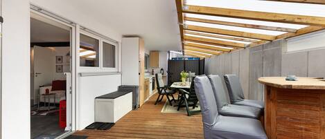 A narrow, covered outdoor space with a wooden floor, a dining table with chairs, and a bar area.