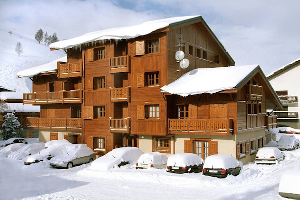 A multi-story wooden building with balconies, a ski lift in the background, and snow-covered cars in front.