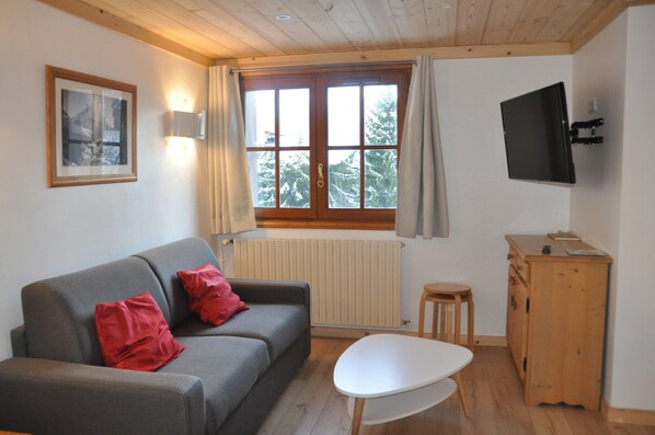 Ein gemütliches Zimmer mit grauem Sofa, einem kleinen weißen Tisch, einem Holzschrank, einem Fernseher und einem Fenster mit Vorhängen.