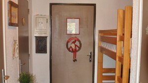 Un pasillo estrecho con techo de madera, una puerta adornada con un adorno, una pequeña mesa con una planta y una cama litera a la derecha.