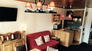 Un espacio habitable compacto con sofá rojo, armarios de madera, una mesa de comedor con un estampado animal único, kitchenette y un televisor de pantalla plana montado en la pared.