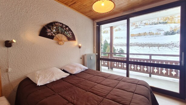 A bedroom with a large bed, a wooden ceiling, a wall-mounted fan, and a view of a snowy landscape through a balcony. - Apt 4/6 ppl with balcony, near the ski station (Les Deux Alpes)