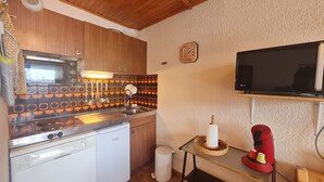 A compact kitchen with wooden cabinets, a microwave, an oven, a dishwasher, a sink, a tiled backsplash, a clock, a television, and a small table with a basket. - Apt 4/6 ppl with balcony, near the ski station (Les Deux Alpes)