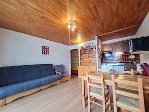 Un salon chaleureux avec un plafond en bois, un canapé bleu, une table à manger avec des chaises et un coin cuisine doté de meubles en bois et d’une cuisinière.