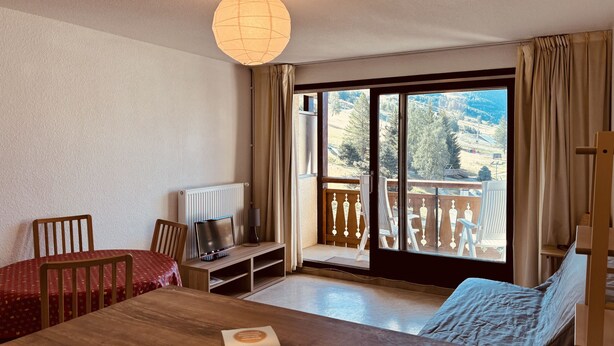 Une pièce comprenant une table à manger, des chaises, un téléviseur posé sur un meuble, un canapé et une vue sur un paysage montagneux, visible à travers une grande porte-fenêtre coulissante.