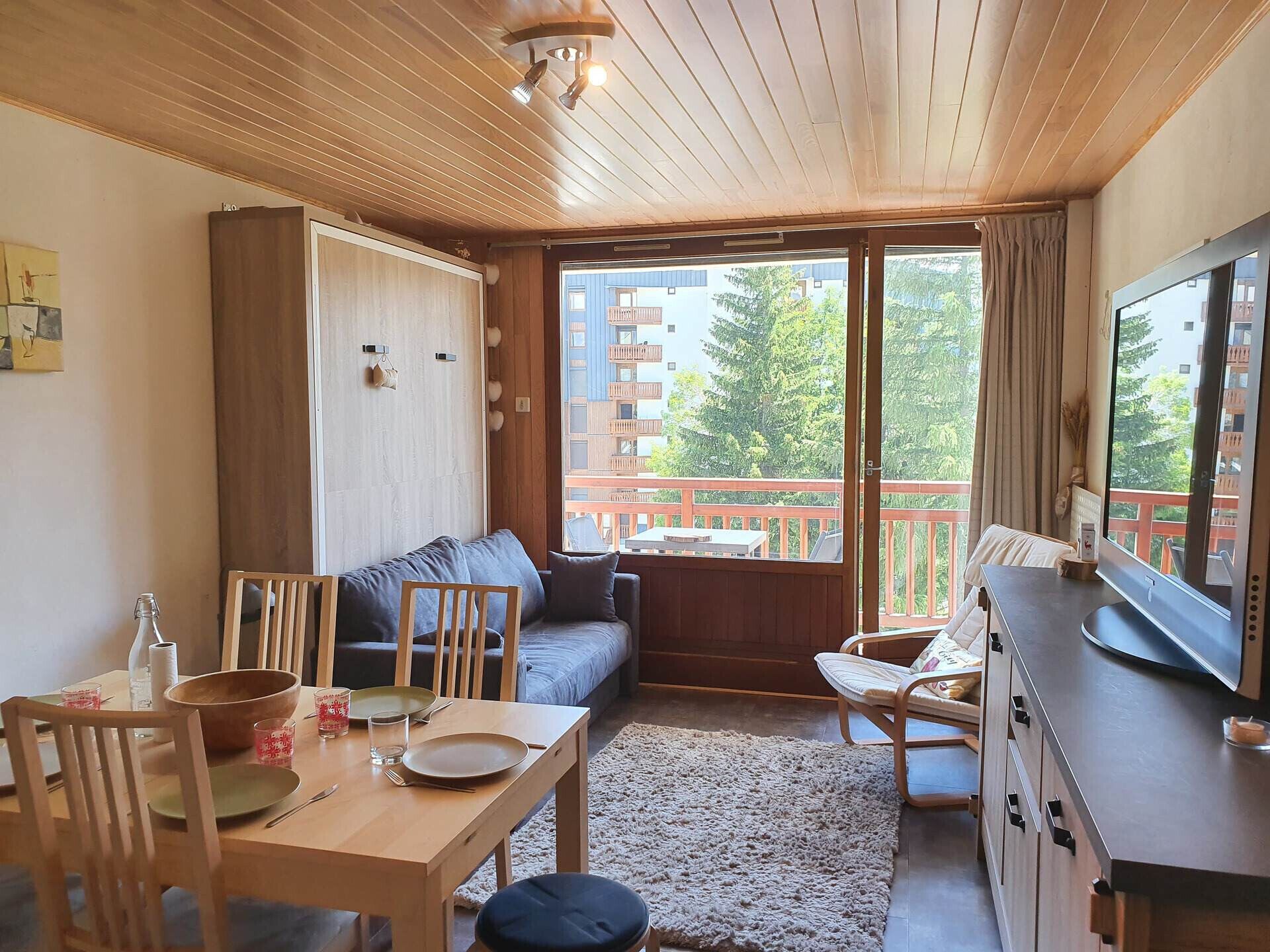 A cozy room with a wooden ceiling, a sofa, a dining table with chairs, a TV, and a balcony with a view of buildings and trees.