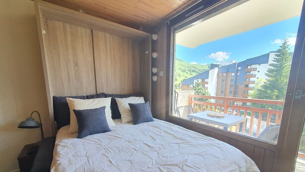 Un dormitorio con una cama grande, cabecera de madera, mesita de noche, lámpara y un balcón con vistas a edificios y árboles.