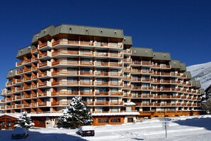 A multi-story hotel with balconies, a ski slope in the background, and a snow-covered ground.