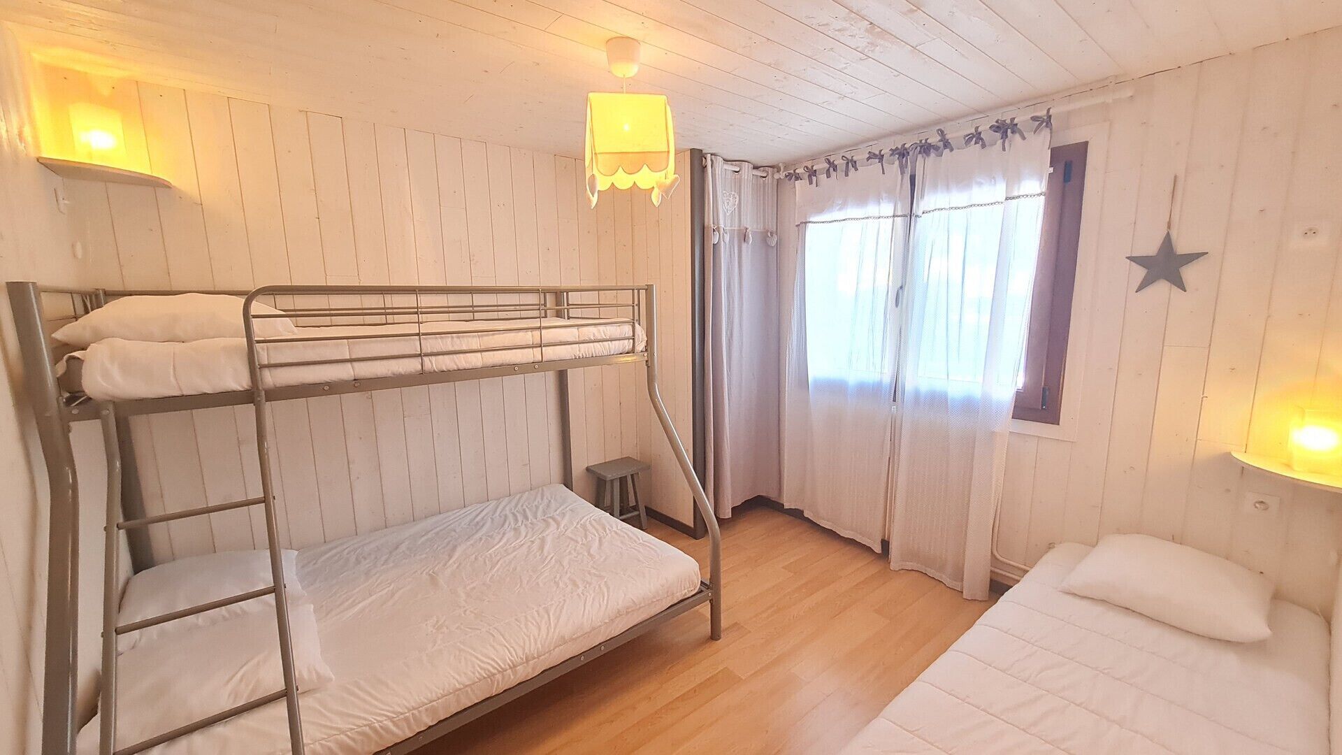 A bunk bed room with a small stool, a window with curtains, and a star decoration on the wall.